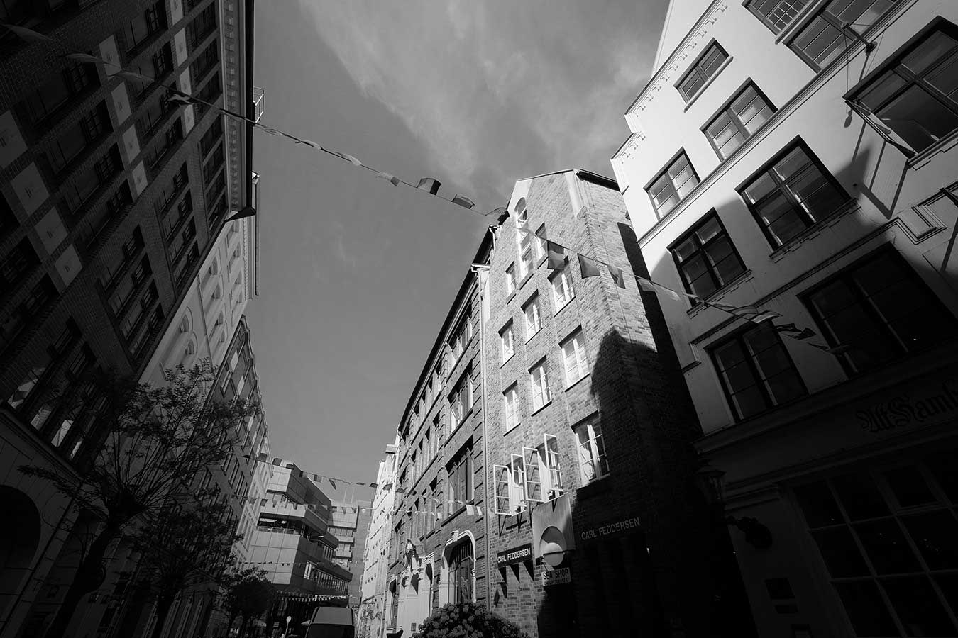 Black and white photo in Hamburg, Germany. Several rows of small rectangular flags are hung between buildings facing each other along a narrow street at sunset.
